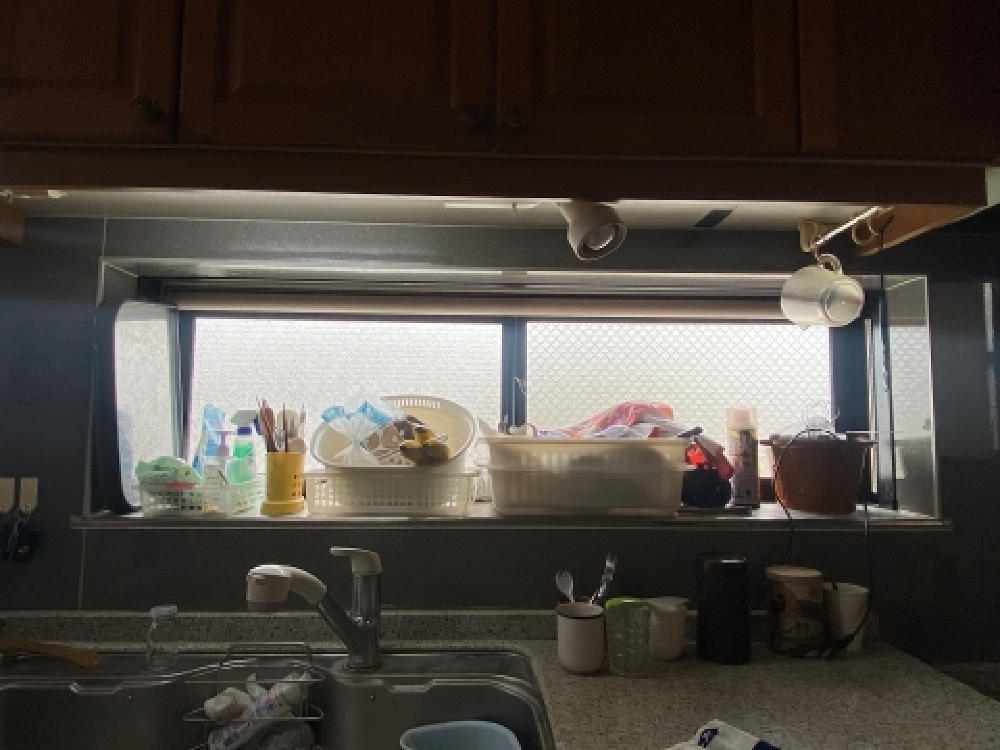 北方町H様邸のリフォーム前キッチン。シンク前の横長窓に食器や洗剤が並び、吊戸棚で採光が遮られている様子。